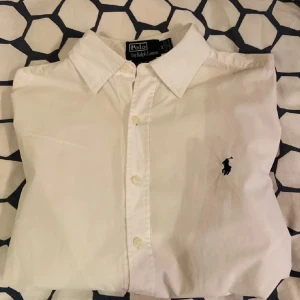 Vit skjorta från Ralph Lauren - Klassisk vit skjorta från Ralph Lauren med broderad logotyp på bröstet. Skjortan har en normal passform och är tillverkad i bomull. Perfekt för en stilren look.