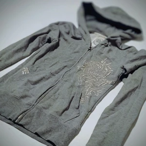 Grå hoodie med drakbroderi - Snygg grå hoodie med dragkedja och ett coolt drakbroderi på framsidan. Perfekt för en avslappnad stil med en touch av edge. Huvan ger extra mysfaktor.