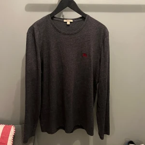 Mörkgrå långärmad tröja från Burberry - Säljer en stilren mörkgrå långärmad tröja från Burberry med en liten broderad logotyp på bröstet. Perfekt för en avslappnad men ändå elegant look.