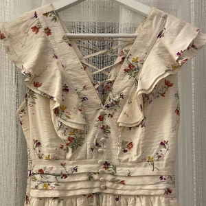 Blommig klänning med volangdetaljer - Charmig blommig klänning i ljus färg med volangdetaljer och snörning i ryggen. Klänningen har en v-ringad design och knappar framtill. Ser ut som ny, super gullig och fin klänning✨✨