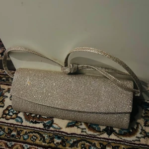 Glittrig kuvertväska från Dorothy Perkins - Snygg glittrig kuvertväska från Dorothy Perkins i en elegant guldton. Väskan har en praktisk axelrem och stängs med en magnetknapp. Perfekt för att ge en touch av glamour till din outfit.