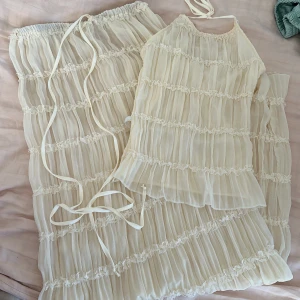 Beige tvådelad klänning - tvådelad klänning i beige med volanger och spetsdetaljer. Toppen är ärmlös med knytband och kjolen har en elastisk midja för bekväm passform. 