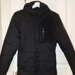 Svart jacka från Sail Racing - Säljer en svart jacka från Sail Racing i nyskick. Jackan har en dragkedja framtill och en praktisk huva. Perfekt för kalla vinterdagar med sin varma och bekväma passform. Den har långa ärmar och en stilren design som passar till det mesta. Perfekt för både vardag och utomhusaktiviteter.