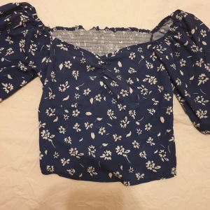 Topp - Blå topp med vita blommor. Toppen har en elastisk kant vid halsen och är något kortare i modellen. Ärmarna är tre fjärdedels långa och har en vid modell. Offshoulder säljs för 50