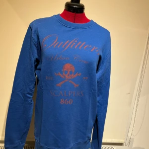 Blå tröja från Scalpers - Säljer en cool blå tröja från Scalpers med tryck av en dödskalle och texten 'Outfitters Urban Crew'. Tröjan är i 100% bomull och har långa ärmar. Perfekt för en avslappnad stil. Tillverkad i Portugal.