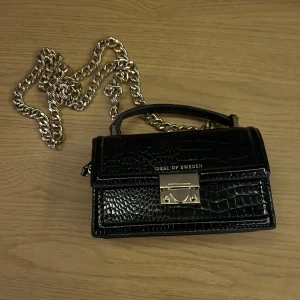 Svart handväska från Ideal of Sweden - Snygg svart handväska i krokodilmönstrat skinn från Ideal of Sweden. Den har en elegant kedja i guld och ett stilrent lås framtill. Perfekt för en utekväll eller fest! 🖤