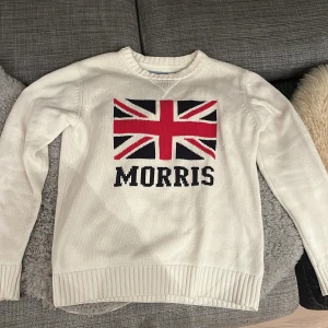 Vit tröja från Morris med Union Jack - Säljer en snygg vit tröja från Morris i storlek medium. Den har ett stort Union Jack-motiv och texten 'Morris' på framsidan. Tröjan är i bra skick och perfekt för både höst och vår. Den är ribbad vid ärmslut och nederkant för en skön passform.