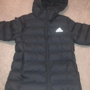 Svart dunjacka från Adidas - Säljer en svart dunjacka från Adidas i nytt skick. Jackan har en dragkedja framtill och en mysig huva. Perfekt för kalla vinterdagar. Adidas-loggan syns tydligt på bröstet. Jackan är både stilren och funktionell, idealisk för vinterns alla äventyr. Köpte jackan för 1299 säljer den för 650