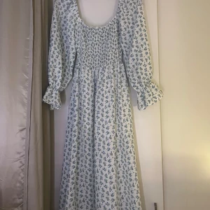 Blommig långklänning med puffärmar - Superfin långklänning i vitt med blått blommönster. Den har puffiga långärmar och en smockad överdel som ger en skön passform. Perfekt för vår och sommar, både till vardags och fest. Klänningen är i ett lätt och luftigt material.
