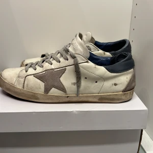 Golden Goose - Säljer ett par vita sneakers med grå stjärndetalj på sidan. Skorna har en cool, sliten look dock lite slitna i hälen (sista bilden). Perfekta för en avslappnad stil och passar till det mesta. De har snörning och är i rätt så bra skick trots den avsiktliga slitna designen.