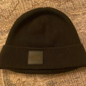 Svart ribbad mössa från Boss - Säljer en stilren svart ribbad mössa från Boss. Den är perfekt för kalla dagar och har en snygg logga framtill. Mössan är i mycket bra skick och passar både till vardags och mer uppklädda tillfällen. Ett måste för garderoben i vinter! 🖤