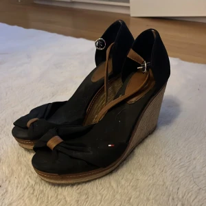 Svarta kilklackar med rosett - Snygga svarta kilklackar från Tommy Hilfiger med en elegant rosett framtill. De har en bekväm kilklack i trälook och justerbara remmar i brunt. Perfekta för sommarens fester eller en dag på stan. Använda men i bra skick.