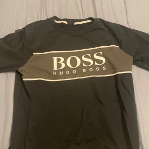 Svart t-shirt från Hugo Boss  - Snygg svart t-shirt från Hugo Boss med en bred vit rand över bröstet där märket står i stora bokstäver. Perfekt för en stilren och avslappnad look. T-shirten är kortärmad och i bra skick. Passar både till vardags och fest!
