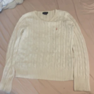 Vit stickad tröja från Ralph Lauren - Säljer en superfin vit stickad tröja från Ralph Lauren. Den har ett klassiskt kabelstickat mönster och en liten broderad logga på bröstet. Perfekt för både höst och vinter, och den är i mycket bra skick. Passar perfekt till jeans eller kjol för en stilren look!