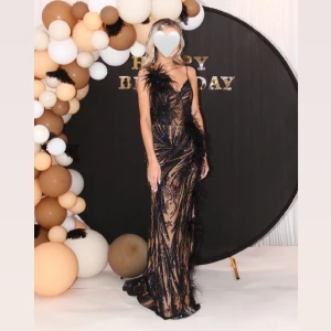 Svart långklänning med fjäderdetaljer - Elegant svart långklänning med fjäderdetaljer och smala axelband. Klänningen har en figurnära passform, men en slits på sidan av benet och är perfekt för festliga tillfällen. Den har en glamorös känsla med sina glittriga inslag och fjädrar som ger en unik touch.                                                                                Klänningen är i nyskick, endast använd 1 gång. I storlek xs/34.