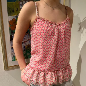 Blommig linne i rosa och vit - Säljer ett sött linne med smala axelband och volangdetalj i nederkant. Linnet har ett blommigt mönster i rosa och vitt, perfekt för sommardagar. Det är i mycket bra skick och passar perfekt till jeans eller shorts.