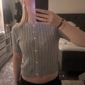 Grå stickad kofta med pärlemorknappar - Säljer en superfin grå stickad kofta med korta ärmar och snygga pärlemorknappar. Perfekt för både vår och höst! Den är croppad och har ett klassiskt flätmönster som ger en stilren look. Passar perfekt till både jeans och kjol. 🩶 