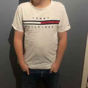 Vit t-shirt från Tommy Hilfiger - Säljer en snygg vit t-shirt från Tommy Hilfiger i ekologisk bomull. Den har en klassisk design med logotypen tryckt på framsidan i svart och rött. T-shirten är kortärmad och passar perfekt till vardags eller en avslappnad dag ute. Skön och stilren, ett måste i garderoben! PRISET ÄR EJ SKRIVET I STEN!!!