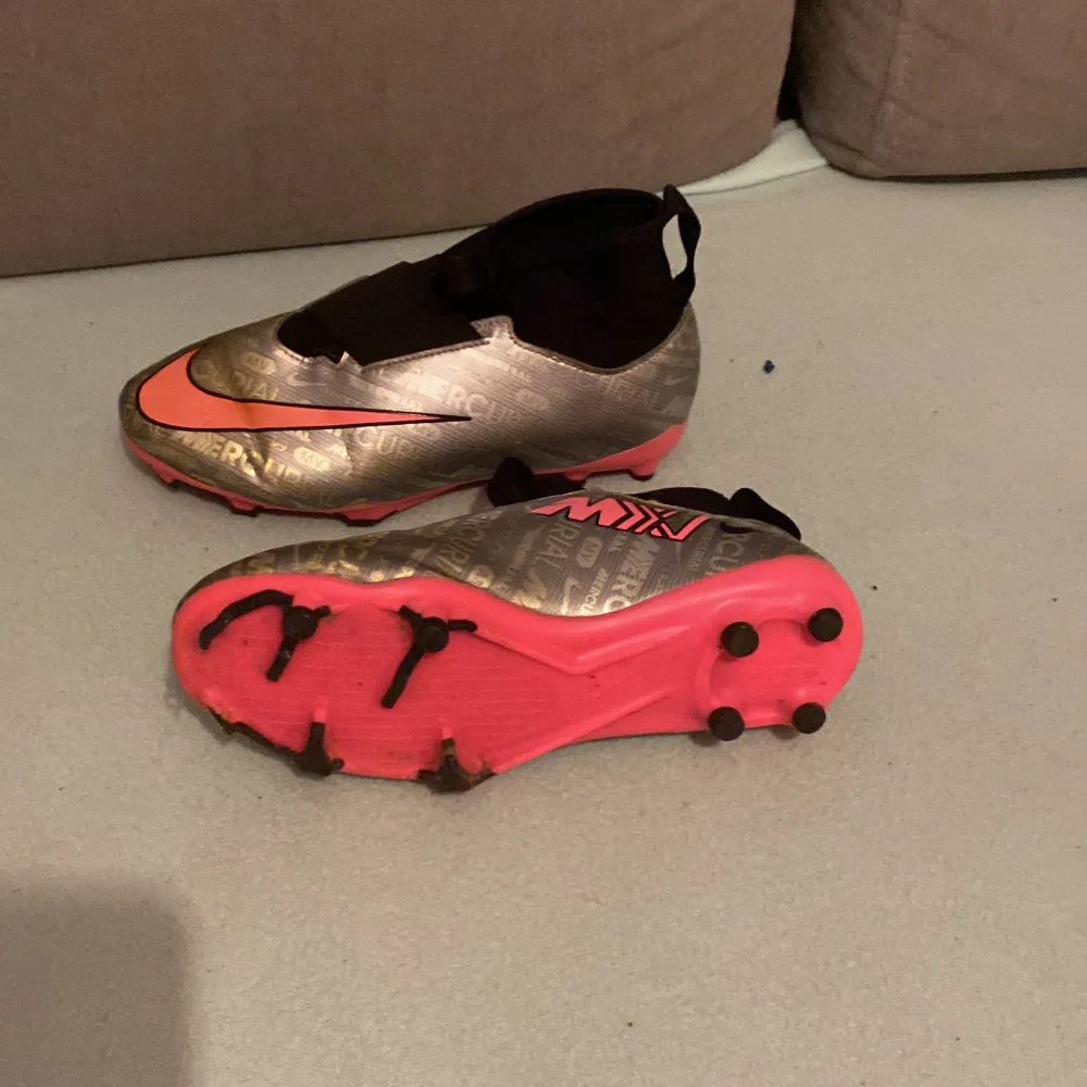 Säljer ett par Nike fotbollsskor i silver med rosa detaljer. Skorna har en cool mönstrad yta och är designade för att ge bra grepp på planen. De har en strumpliknande passform för extra komfort och stil. Perfekta för fotbollsträning eller match!. Kengät.