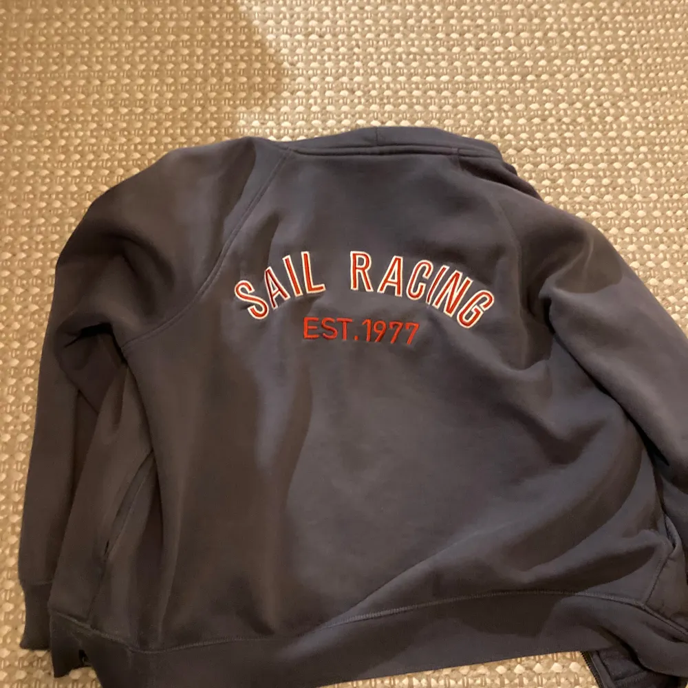 Säljer en snygg mörkblå hoodie från Sail Racing med dragkedja och huva. Den har texten 'SAIL RACING EST. 1977' broderad på ryggen. Perfekt för höst och vår, och passar mycket. Den är i bra skick och superbekväm! Längst ut på armarna syns tecken på användning men annars jättefin. Storlek S/M, är 178 på bilden. Möts upp eller skickar! Vill gärna ha 400 för den men inte satt i sten 😁! Checka in profilen för mer och OskarLiljegren på blocket! Löser paketpris. Hoodies.