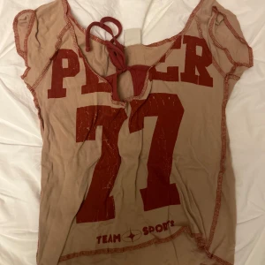 Beige och röd topp med tryck - Säljer en cool beige topp med rött tryck och texten 'TEAM SPORT'. Den har en unik design med knytning i ryggen och korta ärmar. Köp på Urban Outfitters för ca 400kr och knappt använd så mitt pris 200! Passa både S-M🙈💋