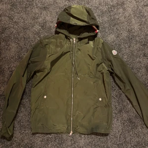 Grön vindjacka från Moncler - Säljer en snygg grön vindjacka från Moncler i mycket bra skick. Jackan har en praktisk huva och dragkedja framtill. Perfekt för höst och vår, med långa ärmar och en stilren design. Moncler-loggan sitter på ärmen. Passar perfekt för både vardag och lättare utomhusaktiviteter.