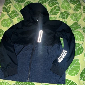 Svart och mörkblå jacka med huva - Säljer en snygg svart och mörkblå jacka med huva. Den har en dragkedja framtill och tryck på ärmen. Perfekt för höst och vår. Jackan har långa ärmar och är i bra skick. Passar både till vardags och lättare utomhusaktiviteter.  12-12 Y 