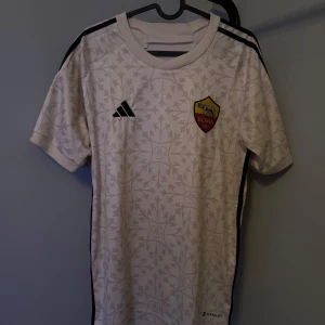 Vit fotbollströja från Adidas - Säljer en vit kortärmad fotbollströja från Adidas med ett subtilt mönster och AS Roma-logga på bröstet. Tröjan har svarta ränder på axlarna och är i bra skick. Perfekt för fotbollsfans eller som träningsplagg! självklart äkta nypris 1199 mitt pris 329