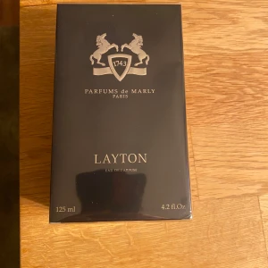 Layton Eau de Parfum från Parfums de Marly - Säljer en  Layton från Parfums de Marly. Flaskan är 125 ml och har en lyxig design med en mörkblå färg och silverdetaljer. Doften är unisex och har varma kryddiga, fräscha kryddiga och vaniljtoner med inslag av trä och citrus. Perfekt för både dag och kväll! Köpt 21 november och är oöppnad, fortfarande inplastad. Onlinekvitto finns. 