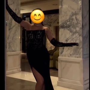 Svart långklänning med spetsdetaljer - Elegant svart långklänning med spetsdetaljer och en slits. Klänningen är axelbandslös och har långa spetsärmar, perfekt för en festlig kväll. Den har en tight passform som framhäver figuren.