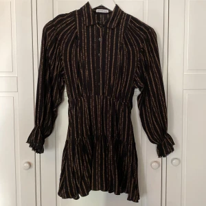 Svart och beige klänning från Designers Remix - Snygg svart klänning med beigerandigt mönster från Designers Remix. Den har långa ärmar med fina detaljer vid ärmsluten och ett knytbälte i midjan för fin böljande passform. Perfekt för höst, vinter och vår!