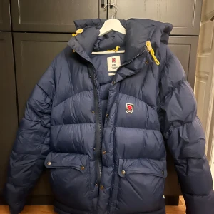 Blå dunjacka från Fjällräven - Säljer en blå dunjacka från Fjällrävens 1974 Expedition Series. Jackan har en mysig huva med gula snören och praktiska fickor med knappar. Perfekt för kalla vinterdagar. Den är i mycket bra skick och redo för nya äventyr! 💙