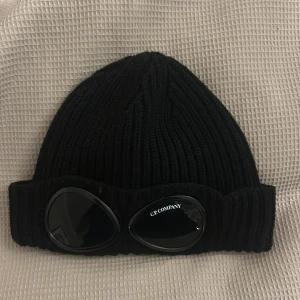 Svart stickad mössa från C.P. Company - Säljer en svart stickad mössa från C.P. Company. Den har en cool ribbad design och inbyggda glasögon på framsidan. Perfekt för att hålla sig varm och stilren under kalla dagar. Passar både till vardags och speciella tillfällen.