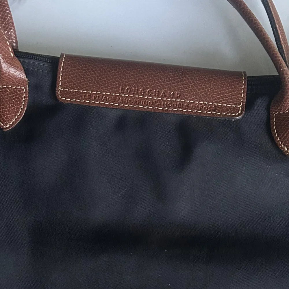 Snygg och praktisk svart axelväska från Longchamp med bruna skinnhandtag och lock. Väskan har en knappdetalj i guld och är perfekt för både vardag och fest. Den är rymlig och stilren, vilket gör den till ett måste i garderoben.Storlek Large. Väskor.