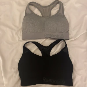 Sport-BH från Reebok i grått och svart - Två sportiga och bekväma sport-BH:ar från Reebok. En i grått och en i svart, båda med brottarrygg och elastiskt band med Reebok-logga. Perfekta för träning och ger bra stöd. Helt nya! 