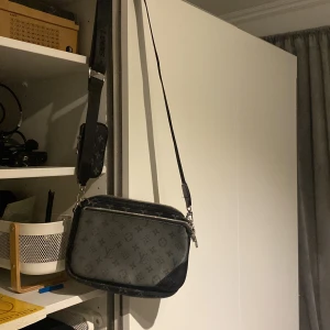 Svart axelväska med monogram - Snygg svart axelväska med monogrammönster. Väskan har en justerbar axelrem och dragkedja för säker förvaring. Perfekt för vardagsbruk eller en kväll ute. Den är i mycket bra skick och har en stilren design som passar till det mesta.