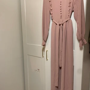 Rosa långklänning från Lovely Line - Säljer en superfin rosa långklänning från Lovely Line. Klänningen har långa ärmar och dekorativa knappar framtill. Den har också volangdetaljer längs med knappraden och ett knytband i midjan för en elegant look. Perfekt för vår och sommar!