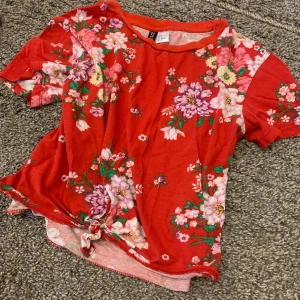 Röd blommig t-shirt från H&M Divided - Säljer en superfin röd t-shirt från H&M Divided med blommigt mönster i rosa och lila. Den har korta ärmar och en knytning framtill som ger en extra touch. Perfekt för sommardagar eller en avslappnad vårlook!