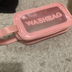 Rosa necessär med dragkedja - Säljer en söt rosa necessär med texten 'WASHBAG' på framsidan. Den har en praktisk dragkedja och ett genomskinligt fönster så du enkelt kan se innehållet. Perfekt för resor eller att ha i badrummet. Handtaget gör den lätt att bära med sig.
