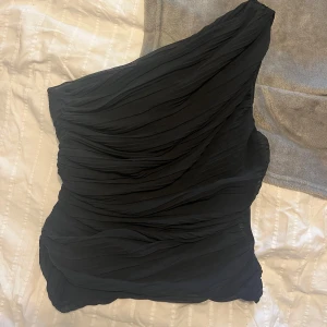 Svart oneshoulder blus från Zara - Snygg svart oneshoulder blus från Zara i ett draperat tyg. Den är i bra skick med en liten reva på innertyget som inte syns när man har på sig den. Använd fåtal gånger. 