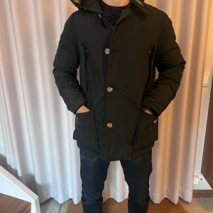 Svart woolrich vinterjacka med huva - Säljer en svart woolrich vinterjacka i mycket bra skick. Den har en huva med pälsdetaljer och stängs med både dragkedja och knappar. Perfekt för kalla vinterdagar! Nypris ca 10000kr. Skick 9/10