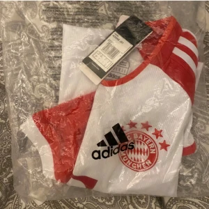 Vit och röd Bayern München fotbollströja från Adidas - Säljer en ny och snygg Bayern München fotbollströja från Adidas. Den är vit med röda detaljer och har korta ärmar. Perfekt för alla fans! Tröjan har klubbens emblem och Adidas-loggan på framsidan. 