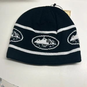 Svart och vit mössa med mönster - Snygg svart mössa med vita ränder och ett coolt mönster. Perfekt för att hålla värmen under kalla dagar. Den är i nytt skick och har en normal passform. Passar perfekt för vintern och hösten.