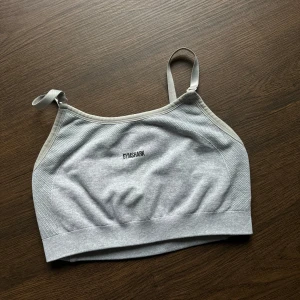 Grå sporttopp från Gymshark - Säljer en grå sporttopp från Gymshark i mycket bra skick. Den är ärmlös med justerbara axelband och har en bekväm passform. Perfekt för träning eller vardagsbruk. Gymshark-loggan syns både fram och bak.