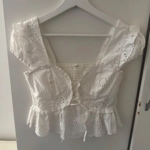 Vit blus med spetsdetaljer - Charmig vit blus med spetsdetaljer och volanger. Den har en smockad midja och korta ärmar med volangkant. Perfekt för en romantisk look.