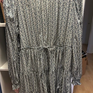 Blommig långklänning med knytband - Säljer en vacker långklänning med blommigt mönster i blått och beige. Klänningen har långa ärmar och ett knytband i midjan för en fin passform. Använd 1 gång 
