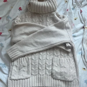 Beige stickad polotröja - Mysig beige stickad polotröja med kabelstickat mönster och praktiska fickor framtill. Perfekt för kyliga dagar med sin höga krage och långa ärmar.