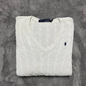 Vit stickad tröja från Ralph Lauren Sport - Säljer en stilren vit stickad tröja från Ralph Lauren Sport med klassiskt kabelstickat mönster och v-ringning. Tröjan har långa ärmar och en liten broderad logotyp på bröstet. Perfekt för en elegant och avslappnad look.