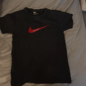 Svart t-shirt med röd Nike-logga - Säljer en svart t-shirt från Nike med den ikoniska röda swoosh-loggan och texten 'Just Do It' på bröstet. Perfekt för en sportig look eller avslappnad stil. Kortärmad och tillverkad i mjukt bomullsmaterial.