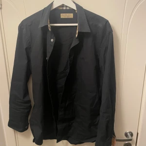 Svart skjorta från Burberry - Snygg svart skjorta från Burberry med klassiskt rutigt mönster på insidan av kragen och ärmsluten. Skjortan har långa ärmar och knappar framtill. Perfekt för en stilren look. Storlek L men passar M lika bra! 